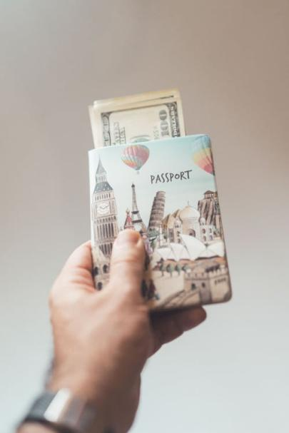 A person holding a passport with dollars inside a creative cover, illustrating the importance of travel preparation and securing necessary documents.